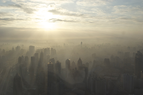 The sun breaks through clouds over a grey city-scape. Smog covers all but the tops of buildings.