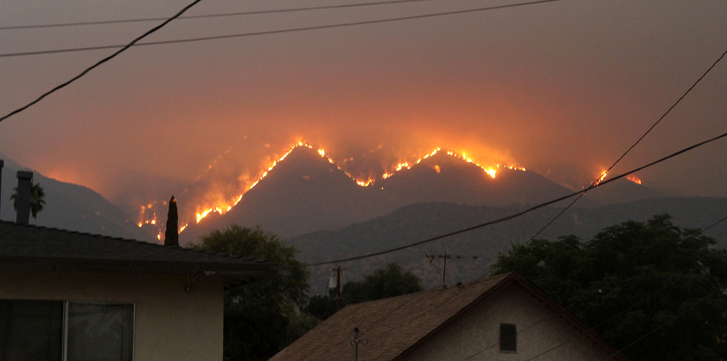 In the background, behind houses, a mountain burns. Orange flames outline the mountain's edges.