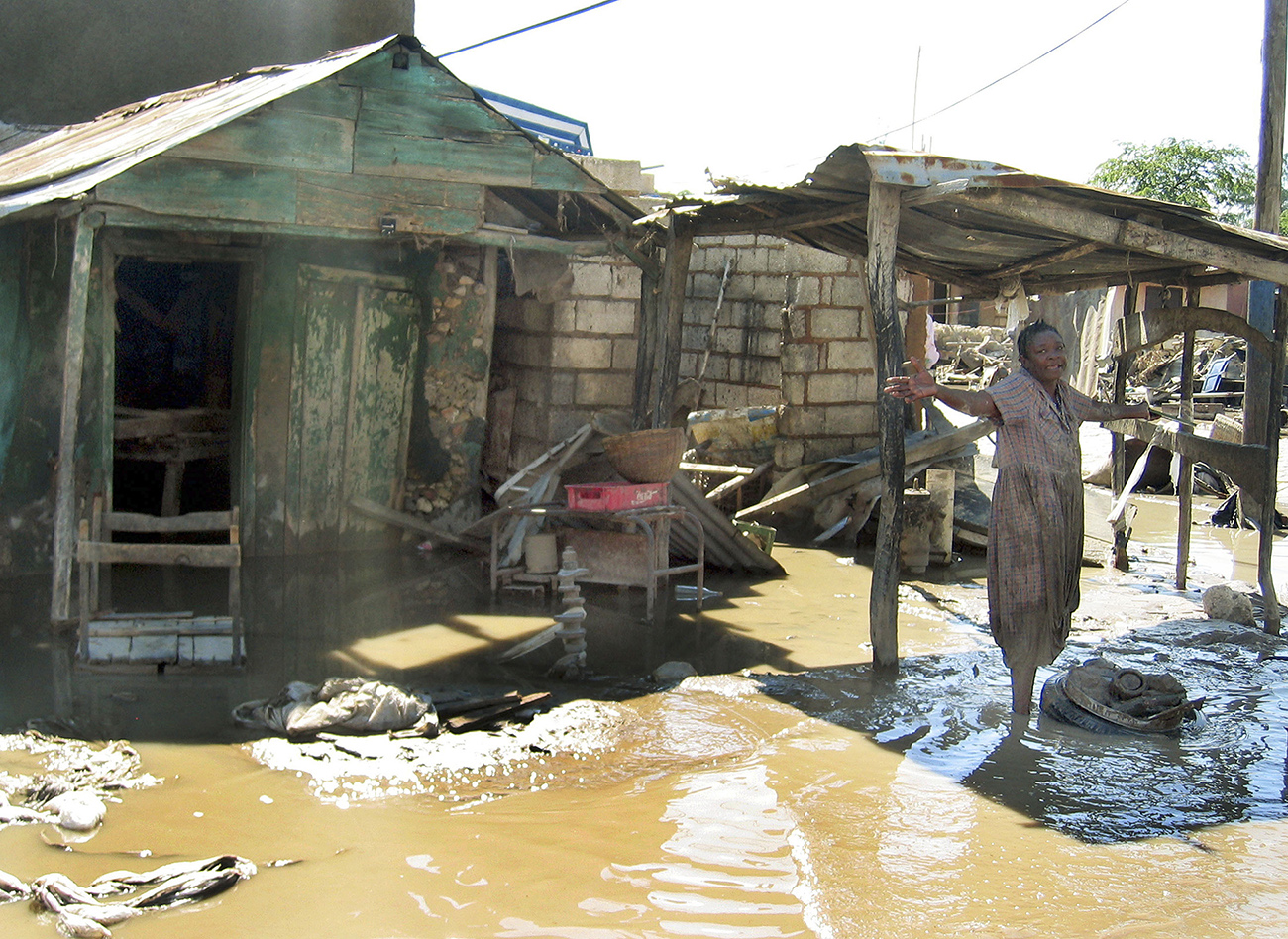A woman stands in a flood in front of a dilapidated house.
