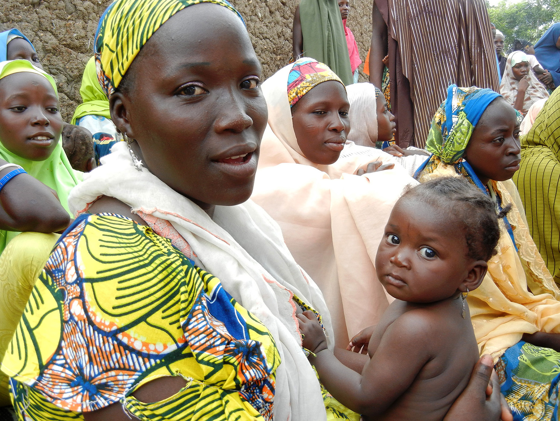 Nigerian woman holding a baby, sitting with a group of other women.